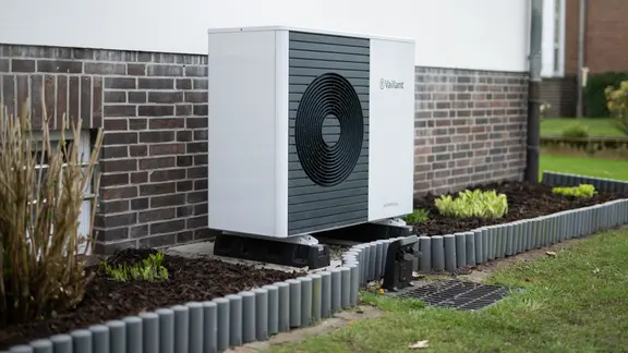 Eine moderne Wärmepumpe des Herstellers Vaillant steht im Garten hinter einem älteren Einfamilienhaus